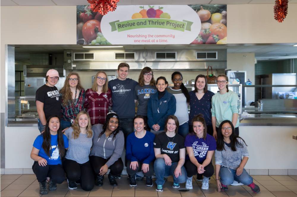 two rows of students in a kitchen area in front of a banner, Revive and Thrive Project, Nourishing the community one meal at a time.